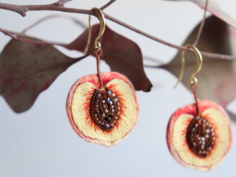 May include: A pair of peach-shaped earrings with embroidered details. The peaches are a light peach color with a darker peach center and brown seed details. The earrings are hanging from gold hooks.