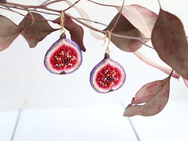 May include: A pair of gold fig earrings with purple and white embroidery. The figs are cut open to reveal a red interior with small, white seeds.