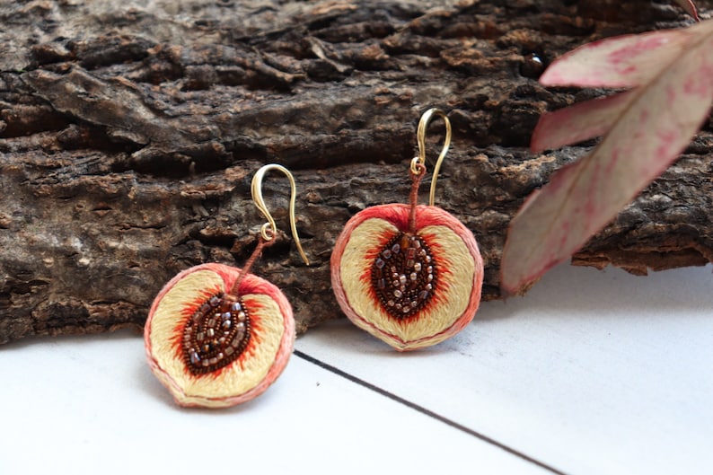 May include: A pair of peach-shaped earrings with gold hooks. The earrings are embroidered with peach slices, with brown seeds and a white center.