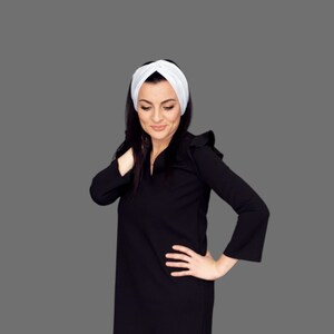 Könnte beinhalten: Eine Frau trägt ein weißes Turban-Haarband und ein schwarzes Kleid mit Rüschen an den Schultern. Sie steht vor einem grauen Hintergrund.