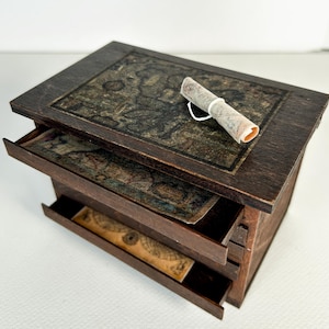 May include: A dark brown wooden chest with multiple pull-out drawers, each featuring a vintage map print on the front. A small, rolled-up paper scroll tied with a white string rests on the top surface of the chest, which also displays a map.