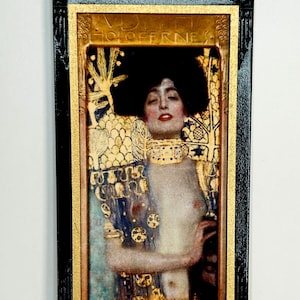 May include: A rectangular framed print featuring a portrait of a person with dark hair, wearing a gold-patterned garment and a wide choker. The artwork includes the text HOLOFERNES at the top, set against a background of gold leaf and intricate patterns.