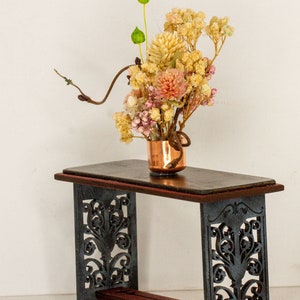 Könnte beinhalten: Eine kleine, kupferfarbene Vase mit einem Trockenblumenarrangement auf einem schwarzen, verzierten Tisch. Die Blumen sind eine Mischung aus getrockneten rosa, weißen und gelben Blüten.