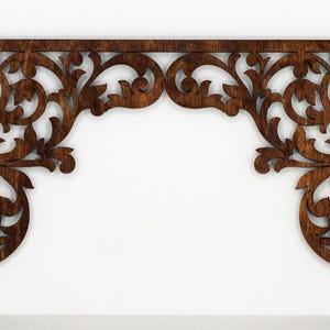 Può includere: Un arco decorativo in legno con una macchia marrone e intricati ritagli floreali. L'arco è in legno e ha un aspetto rustico e vintage.