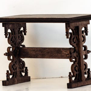 Könnte beinhalten: Ein kleiner, dunkelbrauner Holztisch mit verzierten, geschnitzten Beinen und einer rechteckigen Platte. Der Tisch ist etwa 10 cm lang, 5 cm breit und 7,5 cm hoch. Das Design weist aufwendige Schnörkel und dekorative Elemente auf.