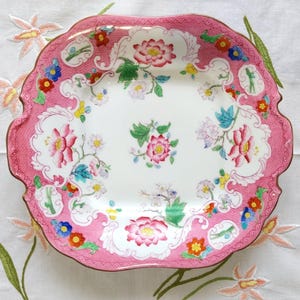 May include: A square porcelain plate with a scalloped edge and a wide pink border featuring a textured pattern. The centre and border are decorated with colourful floral motifs in shades of red, pink, blue, yellow, and green on a white background.
