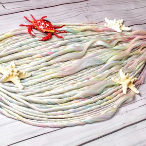 May include: A mermaid-themed hair accessory featuring a bundle of white, pastel-colored dreadlocks. Adorned with a red crab, three starfish, and a purple ribbon. The dreadlocks are arranged on a wooden surface.