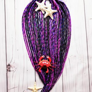 May include: A cluster of purple and black dreadlocks, adorned with two white starfish and a red crab figurine. A sheer, iridescent ribbon is tied at the top. The dreadlocks are arranged against a white, wood-grain background.