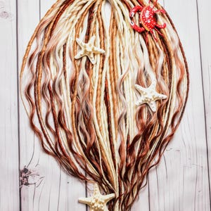May include: A set of dreadlocks in shades of brown, blonde, and red, adorned with white starfish and a red crab figurine. A purple ribbon secures the top of the dreadlocks. The dreadlocks are arranged on a light wood-grain surface.