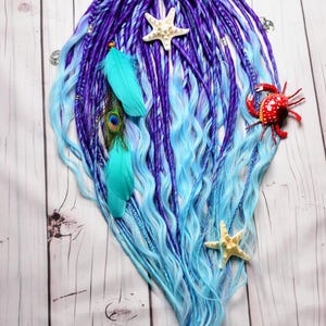 May include: A set of purple and blue dreadlocks with a purple ribbon, adorned with starfish, feathers, and a red crab. The dreadlocks transition from deep purple to a light blue, creating a vibrant, ocean-inspired look.