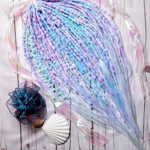 Puede incluir: Un paquete de extensiones de cabello sintético en tonos azules, morados y blancos. El cabello está dispuesto en forma de abanico y está atado con una cinta. El cabello está sobre una superficie de madera blanca.