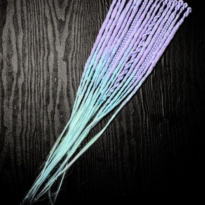 Könnte beinhalten: Ein Bündel synthetischer Haarverlängerungen in einem Farbverlauf von Helllila zu einem sanften Türkis. Die Extensions sind geflochten und auf einer dunklen Holzmaserung angeordnet.