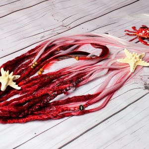 May include: A set of red and white dreadlocks with decorative beads and a starfish. A red crab figurine and a second starfish are also present. The dreadlocks are arranged on a light wood-grain surface.