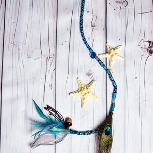 Puede incluir: Una extensión de pelo trenzada azul y verde con una pluma de pavo real, una cuenta azul y una cuenta de madera marrón. La extensión está decorada con dos estrellas de mar.