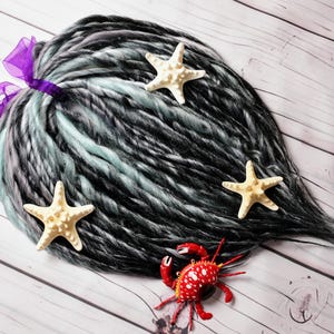 May include: A bundle of black, gray, and teal dreadlocks tied with a purple ribbon. Three beige starfish and a red crab figurine are arranged on a light wood-grain surface, creating a beachy theme.