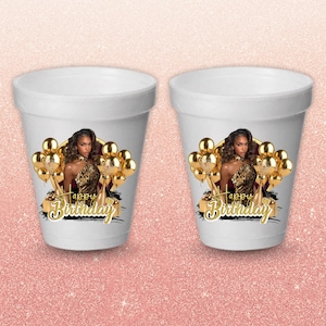May include: Two white foam cups with a printed design. Each cup features a woman in a leopard print top, gold balloons, and the words "Happy Birthday" in gold script. The cups are on a pink glitter background.