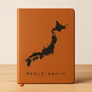 Puede incluir: Un cuaderno de cuero marrón con una silueta negra de Japón. El cuaderno tiene una cinta de marcador y texto japonés en la parte inferior. La cubierta es de color marrón cálido.