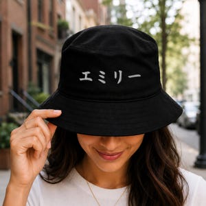 May include: Black bucket hat with white Japanese text. The hat is made of a soft material and has a rounded crown and a downward-sloping brim. The text on the hat reads "Emily".