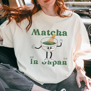 May include: Cream-coloured t-shirt with the words "Matcha in Japan" in green, and Japanese characters. The design features a cartoon bowl of matcha with a whisk, arms, legs, and a smiling face. The t-shirt has a relaxed fit.
