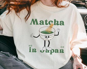 以下が含まれることがあります： 抹茶と日本語で「Matcha in Japan」と書かれたクリーム色のTシャツ。デザインは、泡立て器、腕、脚、笑顔の顔を持つ漫画の抹茶のボウルです。ゆったりとしたフィット感のTシャツです。