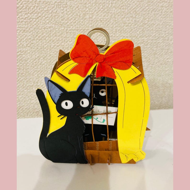 May include: A black cat with blue-tipped ears sits next to a yellow cage with a red bow on top. The cage has a brown base and a metal handle. Inside the cage is a black cat and a small piece of paper.