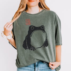 Könnte beinhalten: Ein salbeigrünes Kurzarm-T-Shirt mit einer schwarzen Grafik eines Frosches mit rundem Bauch. Über dem Frosch befindet sich ein kleines rotes quadratisches Stempeldesign. Das T-Shirt hat einen Rundhalsausschnitt und eine lockere Passform aus verwaschenem Stoff.