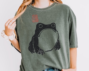 May include: A sage green short-sleeve t-shirt featuring a black graphic of a frog with a round belly. Above the frog is a small red square stamp design. The shirt has a crew neckline and a relaxed fit, made from a washed-out fabric texture.