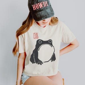 Op de afbeelding: Crèmekleurig T-shirt met een grote zwarte kikkerprint. Het shirt heeft ook een klein rood stempelachtig ontwerp en wordt gecombineerd met een zwarte baseballcap met het woord "HOWDY" in rode letters.
