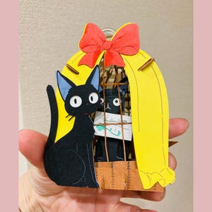 May include: A black cat figurine with blue-tipped ears sits beside a yellow cage with a red bow on top. Inside the cage is another black cat holding a note. The figurine is held in a hand.