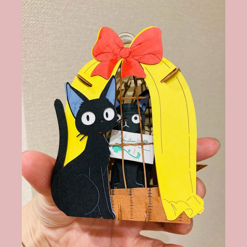 May include: A black cat figurine with blue-tipped ears sits beside a yellow cage with a red bow on top. Inside the cage is another black cat holding a note. The figurine is held in a hand.
