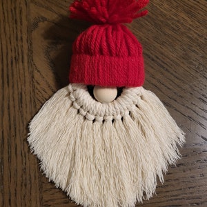 May include: A white yarn gnome ornament with a red knit hat and pom pom. The gnome has a wooden bead nose.