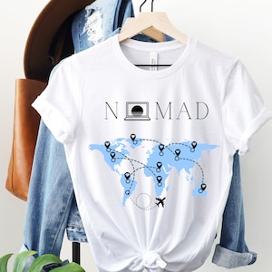 Puede incluir: Camiseta blanca con un mapa del mundo azul y la palabra "NOMAD" impresa en ella. Un icono de computadora port&aacute;til est&aacute; incorporado en la palabra "NOMAD".