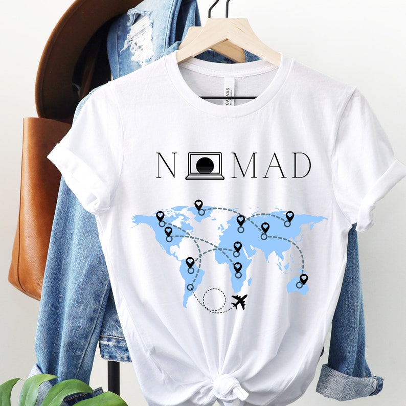 Puede incluir: Camiseta blanca con un mapa del mundo azul y la palabra "NOMAD" impresa en ella. Un icono de computadora port&aacute;til est&aacute; incorporado en la palabra "NOMAD".
