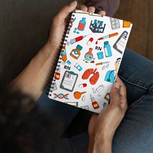 Peut inclure: Un carnet &agrave; spirales blanc avec un motif color&eacute; repr&eacute;sentant des symboles m&eacute;dicaux, notamment des seringues, des pilules et un st&eacute;thoscope. Le carnet porte l'inscription "RN".