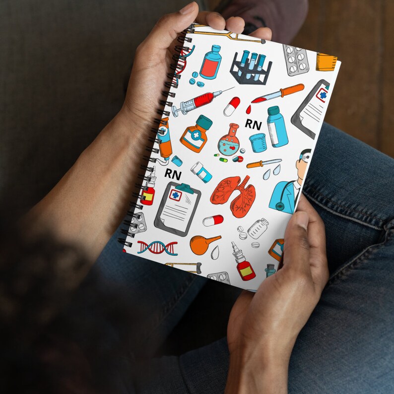 Peut inclure: Un carnet &agrave; spirales blanc avec un motif color&eacute; repr&eacute;sentant des symboles m&eacute;dicaux, notamment des seringues, des pilules et un st&eacute;thoscope. Le carnet porte l'inscription "RN".