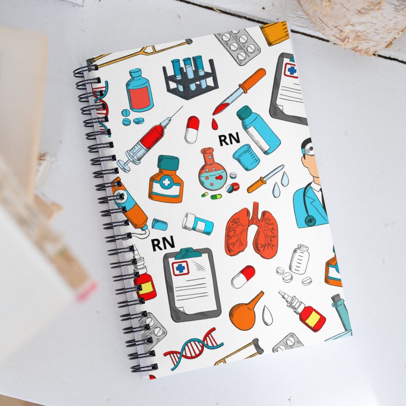 Peut inclure: Un carnet &agrave; spirales blanc avec un motif color&eacute; d'illustrations li&eacute;es &agrave; la m&eacute;decine et aux soins infirmiers. La couverture pr&eacute;sente des seringues, des pilules, des tubes &agrave; essai, un st&eacute;thoscope et le texte "RN".