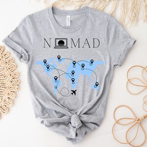 Puede incluir: Camiseta gris con el texto "NOMAD" y un gr&aacute;fico de un mapa del mundo con alfileres y un avi&oacute;n.