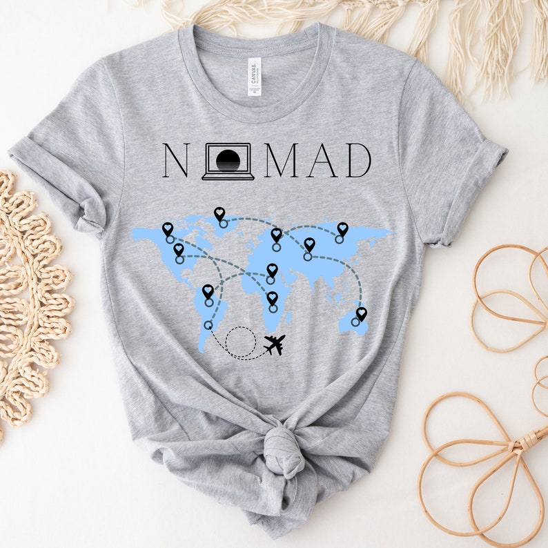 Puede incluir: Camiseta gris con el texto "NOMAD" y un gr&aacute;fico de un mapa del mundo con alfileres y un avi&oacute;n.