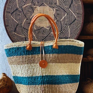 May include: A woven tote bag with horizontal stripes in teal, beige, and tan. The bag has brown leather handles and a matching leather tag. A large, round woven wall hanging is in the background.