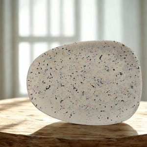 以下が含まれることがあります： 有機的な形状の、まだら模様のオフホワイトのセラミックプレート。プレートには、小さな黒、青、ピンクの斑点のパターンがあります。木製のテーブルの上に置かれ、背景には窓と椅子がぼやけています。