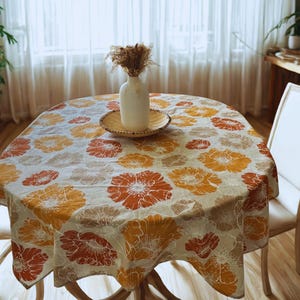 May include: A round dining table set with a floral tablecloth in shades of orange, red, and beige. The table is set with a white vase, a woven plate, and dried flowers. Two white chairs are positioned on either side of the table.