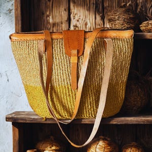May include: A woven straw tote bag with a leather flap closure and shoulder straps. The bag is a mix of yellow and natural straw colors. The bag is sitting on a wooden shelf with other woven items.