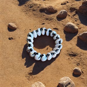 May include: A circular bracelet made of white, oval-shaped beads with black accents. The bracelet is resting on a sandy surface with small rocks scattered around it. The beads have a glossy finish.
