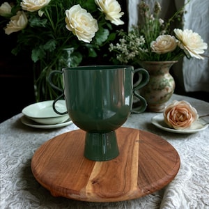 Könnte beinhalten: Eine dunkelgrüne dekorative Vase mit zwei Griffen, die auf einem runden Holztablett steht. Die Vase ist der Mittelpunkt, mit einem Sockel. Weiße Rosen und andere Blumen sind im Hintergrund und schaffen eine natürliche, elegante Ästhetik.