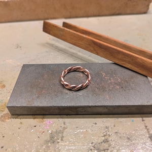 Könnte beinhalten: Ein Kupferring mit einem gedrehten, geflochtenen Design. Das Band besteht aus mehreren miteinander verflochtenen Strängen aus metallischem Kupferdraht, die ein durchgehendes, gleichmäßiges Muster um den gesamten Umfang des Rings bilden.
