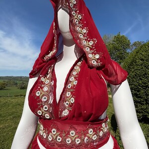 May include: A red, sheer, embroidered dress with a matching head scarf. The dress has a deep V-neckline and a fitted bodice. The skirt is flowing and has a gathered waist. The embroidery is gold and silver and features floral and geometric patterns.