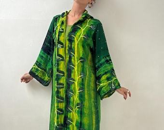May include: A long, flowing green robe with a botanical design in shades of yellow and black. The robe features long sleeves and a hood. Small, sparkling embellishments are scattered across the fabric, adding a touch of shimmer. The robe is worn open, revealing a green top underneath.