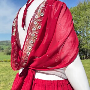 May include: A red chiffon saree with a gold and silver embroidered border. The saree is draped over a white mannequin and is tied at the waist.