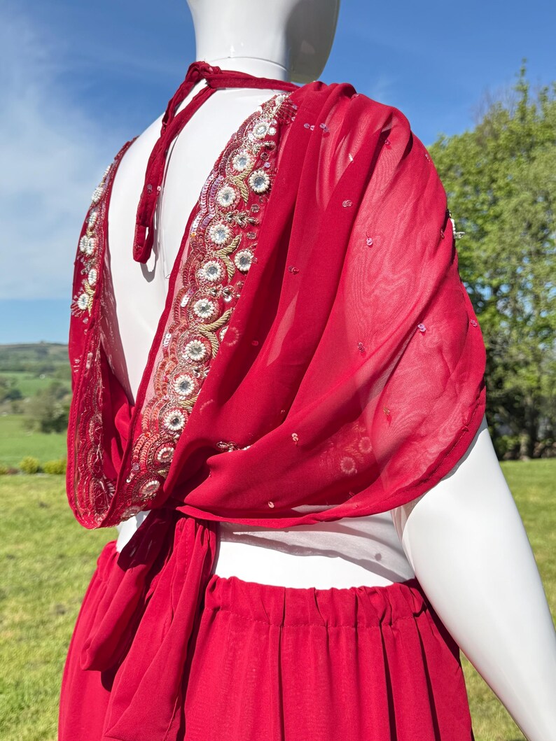 May include: A red chiffon saree with a gold and silver embroidered border. The saree is draped over a white mannequin and is tied at the waist.