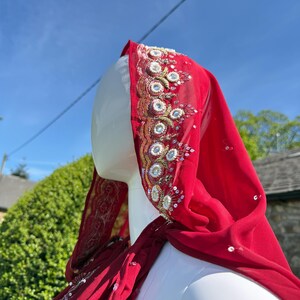 May include: A red sheer cape with gold and silver embroidery. The cape has a hood and is decorated with sequins and beads.